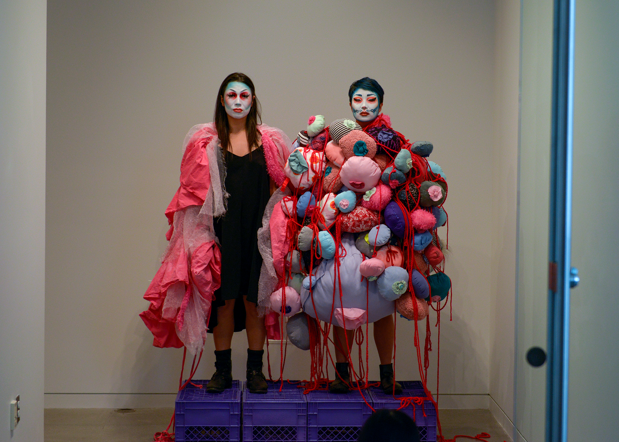 SKIM and Maiden China stand on blue crates in bold coloured makeup covering their faces, looking fiercely into the camera.