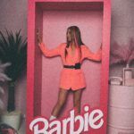 Pink convocation, unnecessary vent sesh, and opposite day? Portrait of a woman posing in a large Barbie box. She is surrounded by pink items, including a rotary phone and ice skates.