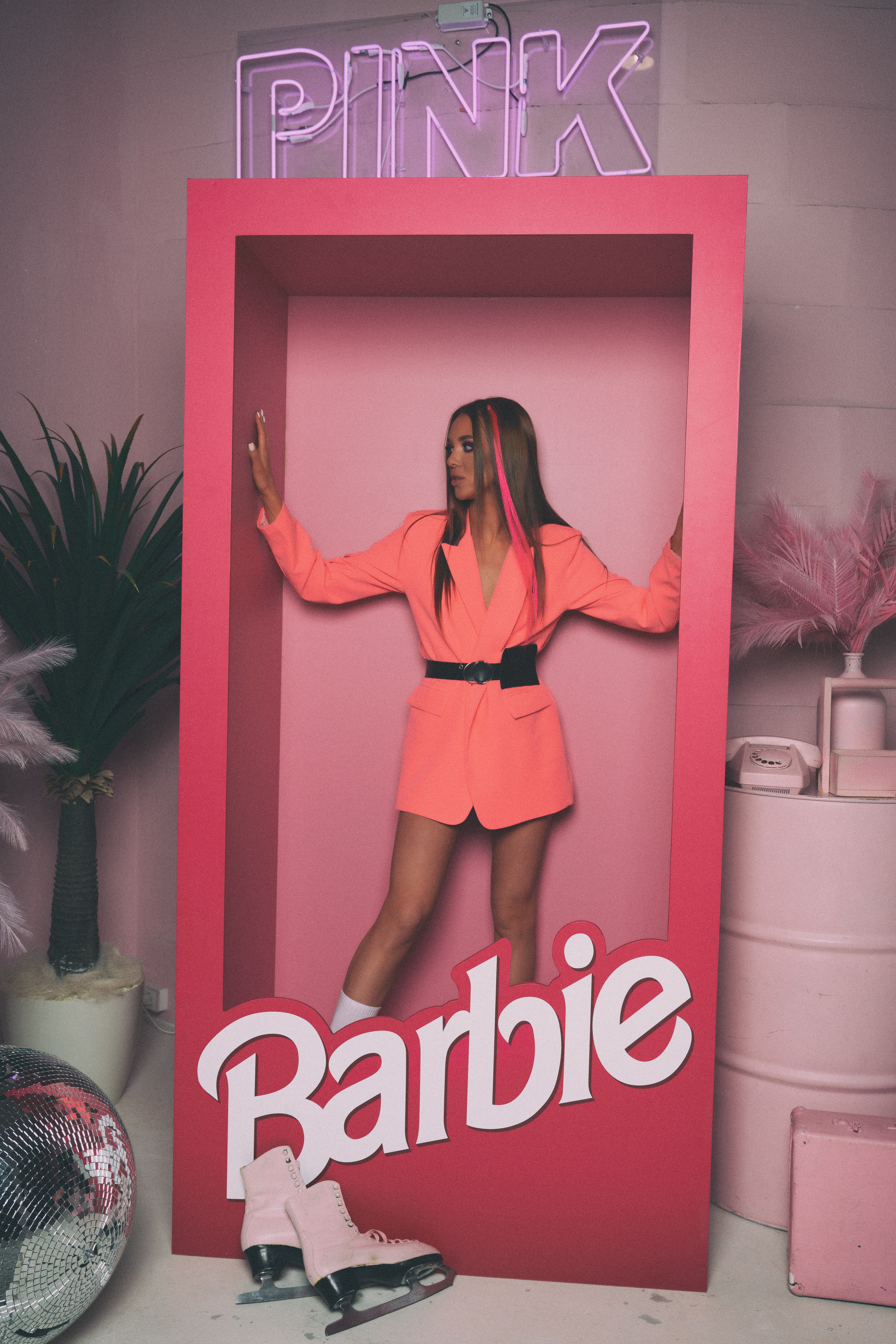 Portrait of a woman posing in a large Barbie box. She is surrounded by pink items, including a rotary phone and ice skates.