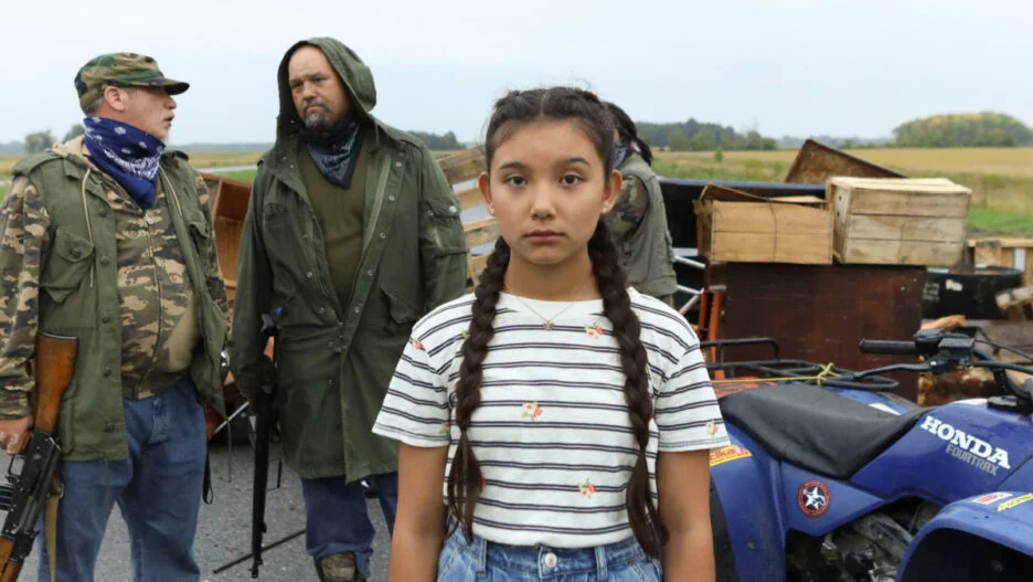 Beans film still of a young Indigenous girl standing solemnly in front of two Indigenous men dressed in camouflage with guns. The background shows an expansive field, a motorcycle. The girl has two braids and is wearing a striped shirt.