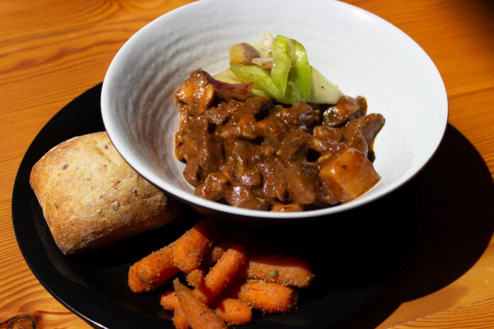 IMG_3782 A large white bowl with bison bourguignon, a stew with chunks of meat, carrot, and vegetable garnish. On the side, on a plate, is a loaf of bread and carrots.
