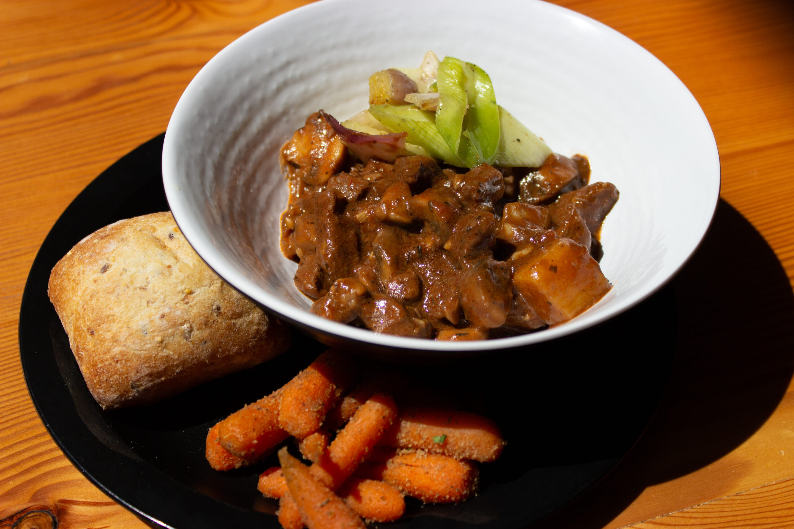 A large white bowl with bison bourguignon, a stew with chunks of meat, carrot, and vegetable garnish. On the side, on a plate, is a loaf of bread and carrots.