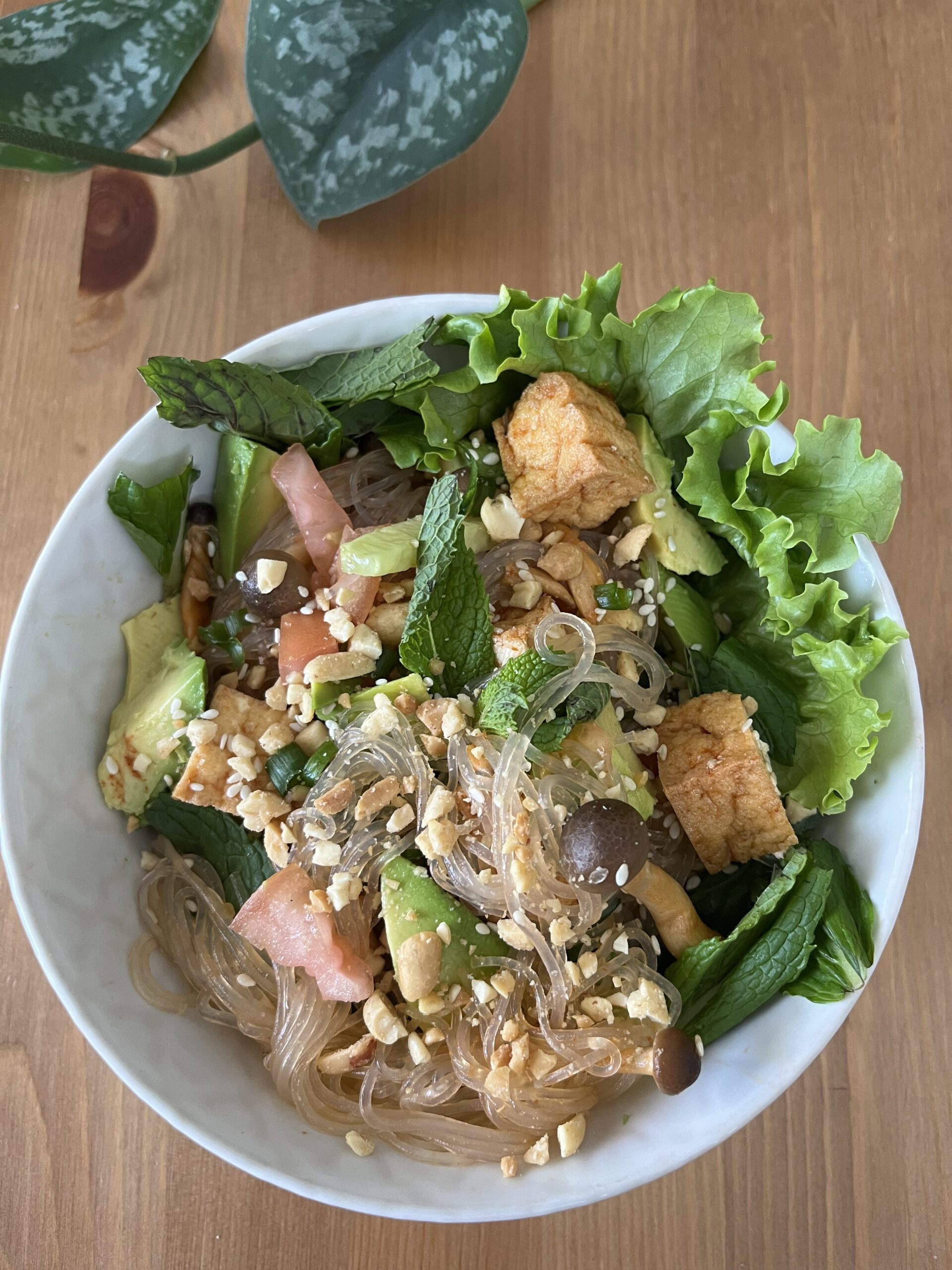 A photo of a bowl of noodle salad on a wooden table. The bowl has lettuce, tofu, mushrooms, and more vegetables.