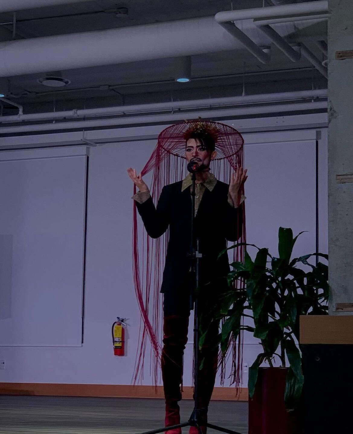 King Fisher performing a drag set, wearing bold makeup and a sharp collared shirt with a blazer. He is in front of a microphone sitting under a hanging string ornament.