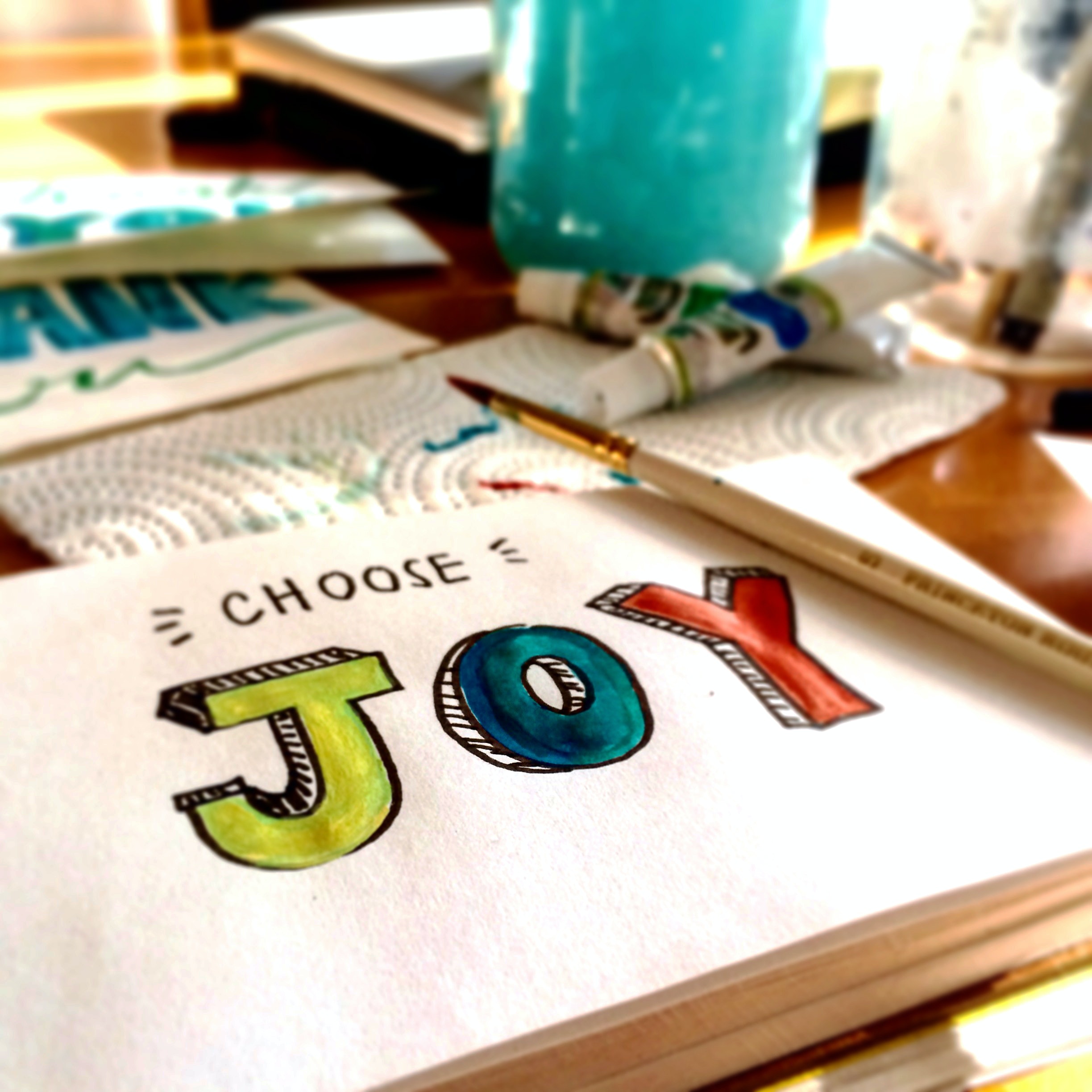White paper with “choose joy” painted on it. The paper is surrounded by art supplies.