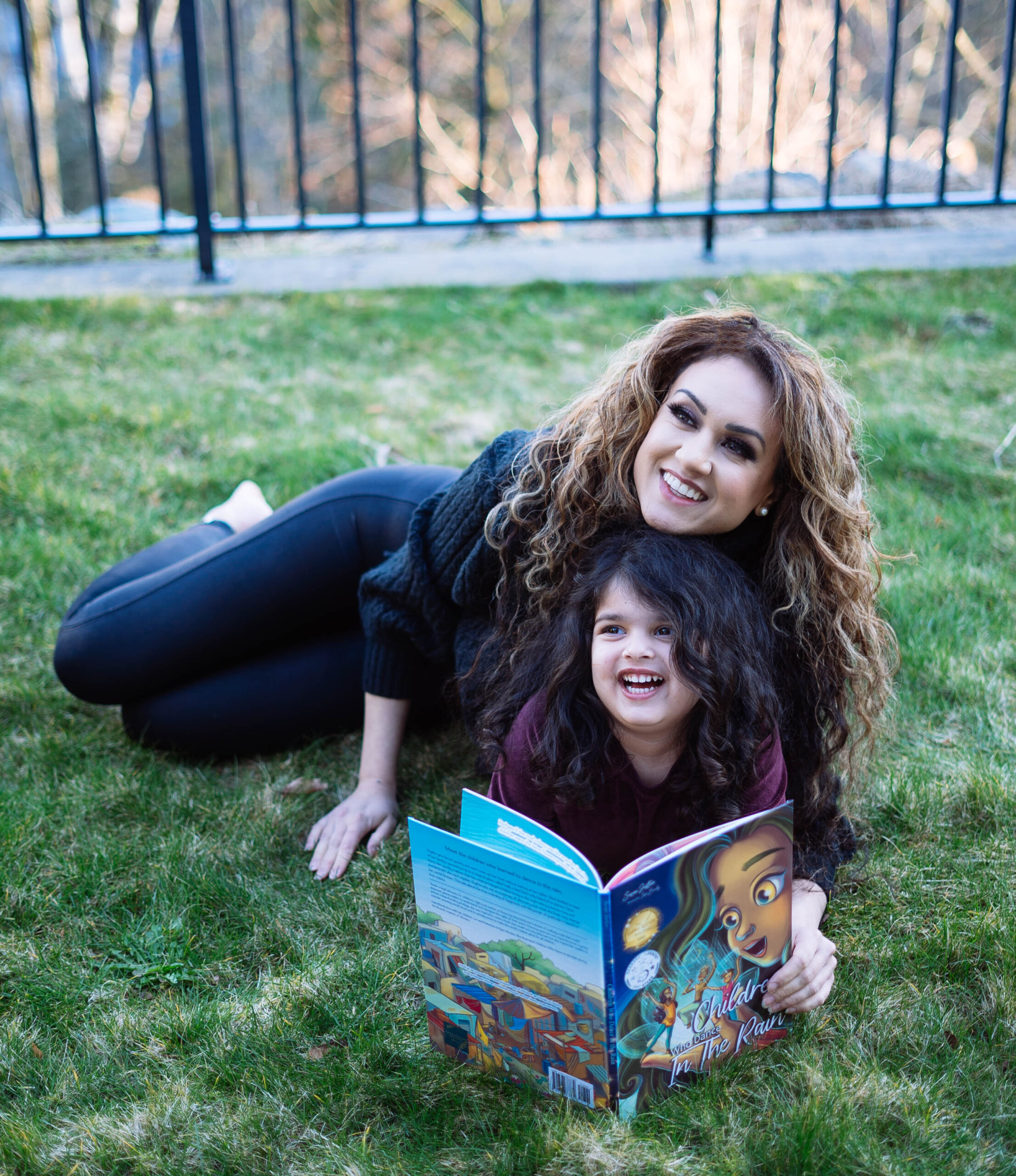 Susan Justice and child reading a the Children in the Rain book on a grass lawn with wide smiles.