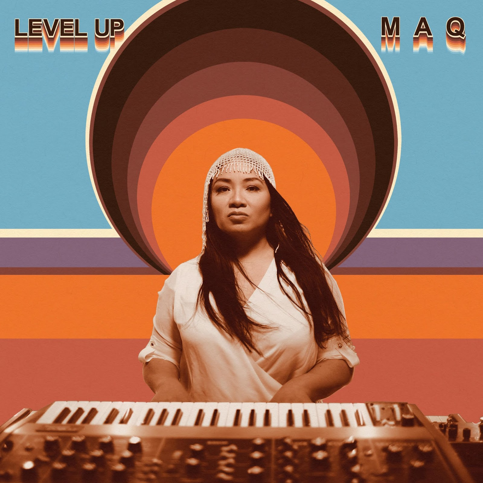 Album cover featuring a photo of Mary Ancheta behind a psychedelic background with a spiral with stripes, warm orange, blue, and purple. Ancheta is playing a keyboard and looking fiercely into the camera. She had long black hair, wearing a white headpiece and silky shirt.