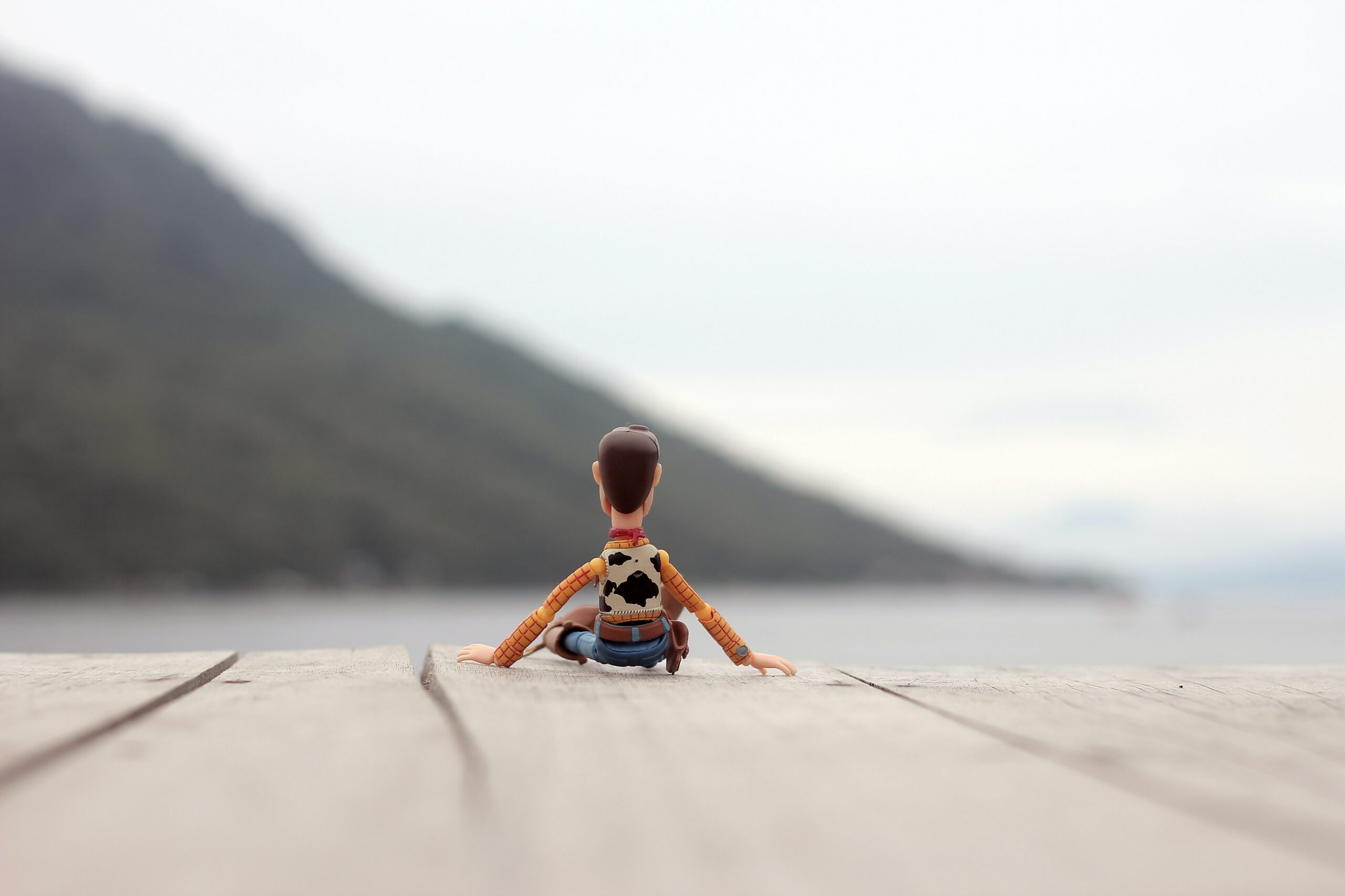 A toy figurine of the charachter Woody from the Pixar movie Toy Story sitting, in the horizon a mountain and the sky can be seen