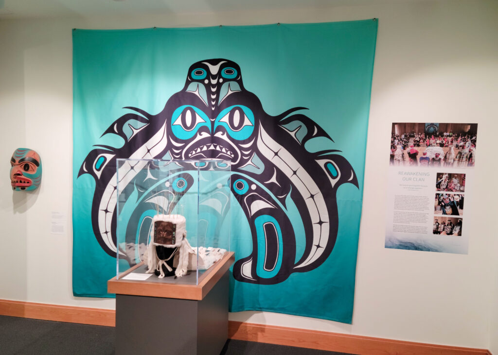 A sculpture of an abstract creature, its face carved in metal, whiskers sticking out from its head, and a white fur coat covering its head and falling back. It’s in a metal case and placed in front of a turquoise-blue fabric hanging on the wall with a Haida-style abstract painting.