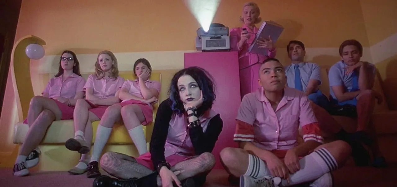A film still featuring a group of bored-looking teens sitting on a couch looking up at a projected video. The projector is shown pointed towards the camera and an older woman is clicking the remote. The girls are dressed in pink button-ups and skirts, and the boys are dressed in blue button ups with ties.