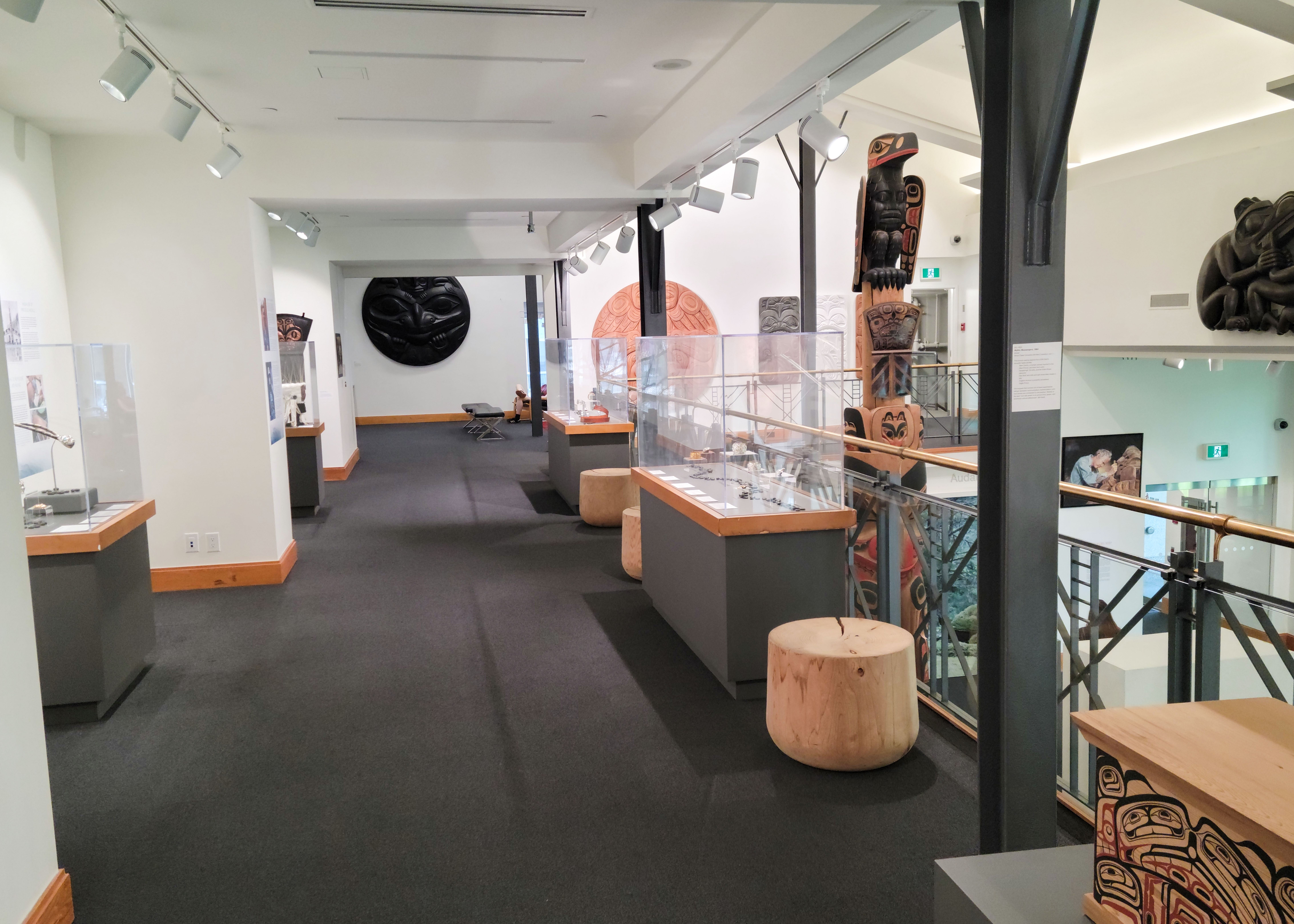 A photo of a corridor on the mezzanine of the Bill Reid Gallery featuring glass cases, Haida-style wood carvings depicting abstract faces and creatures on the wall, and a totem pole emerging from the first floor.