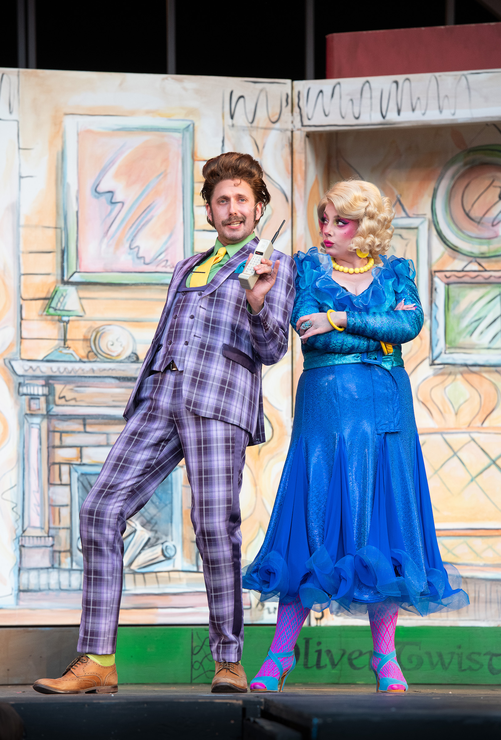 A photo of two characters from Matilda, Mr and Mrs Wormsworth standing on stage in front of an illustrated landscape backdrop. Mrs wormsworth has exaggerated makeup like red cheeks and pale foundation, and fluffy blonde hair. Her arms are crossed and she’s wearing a blue formal dress, staring at Mr. Wormsworth. Mr. Womswoth wears a purple checkered suit, has puffy brown hair and mustache, and has a mischievous look on his face, holding a vintage phone out in one hand.