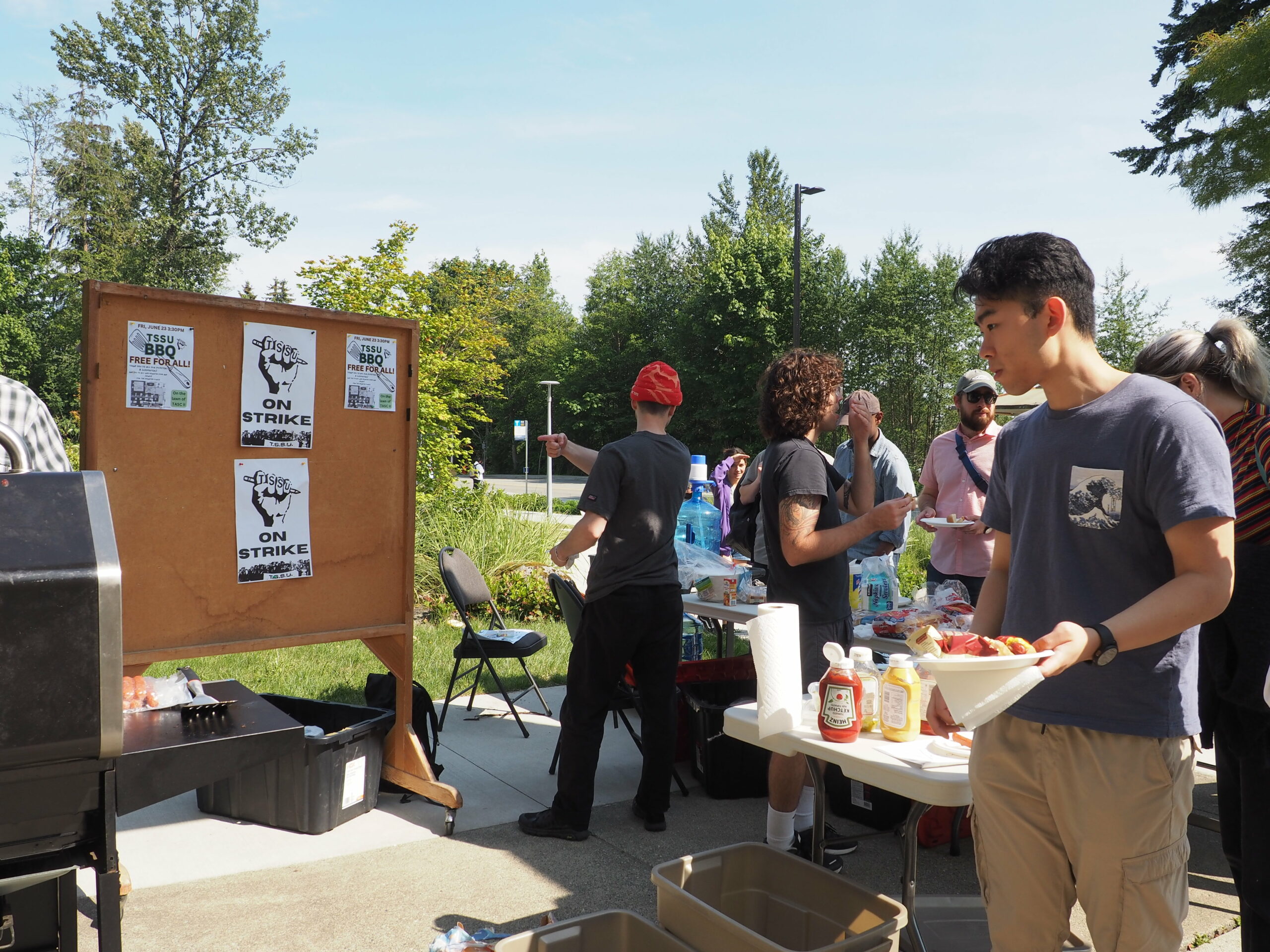 This is a photo taken at the barbeque. People are getting hotdogs from a table. Behind them, there is a cork board that has TSSU signs.