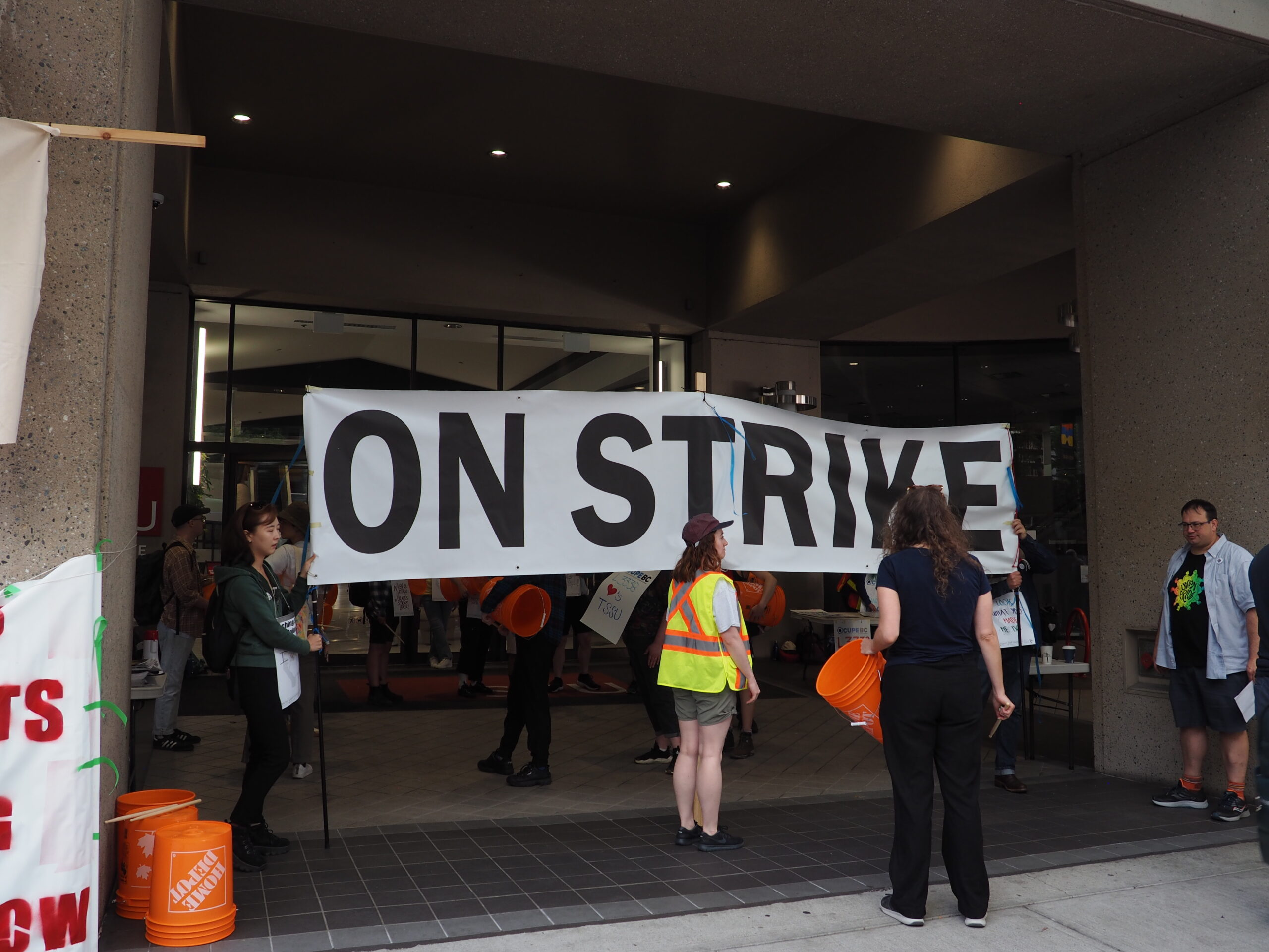 This is a photo of the picket line at SFU Harbour Centre. Members are holding a large banner in front of the doors that reads ON STRIKE