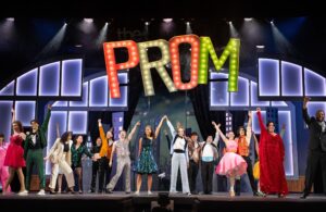 A photo of the cast of The Prom standing on the stage in from of a neon sign that says “prom” Everyone is dressed in sparkle and sequin prom dresses and colourful suits with their fists in the air and joyful expressions.