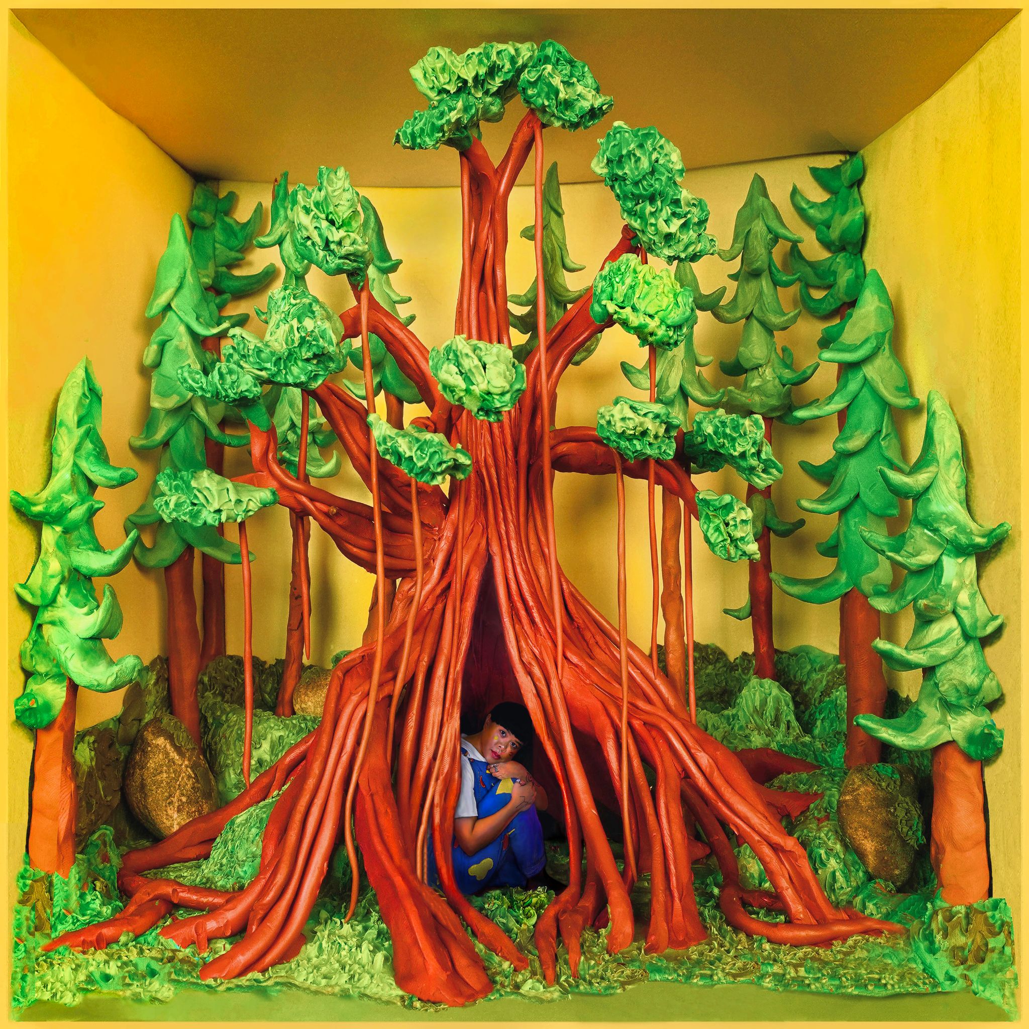 An album cover featuring a clay-molded forest in a yellow box, and a wide, hollow tree stump in the middle, inside which Kimmortal sits holding their legs and looking out playfully. They’re wearing royal blue overalls and have black hair with straight-cut bangs.