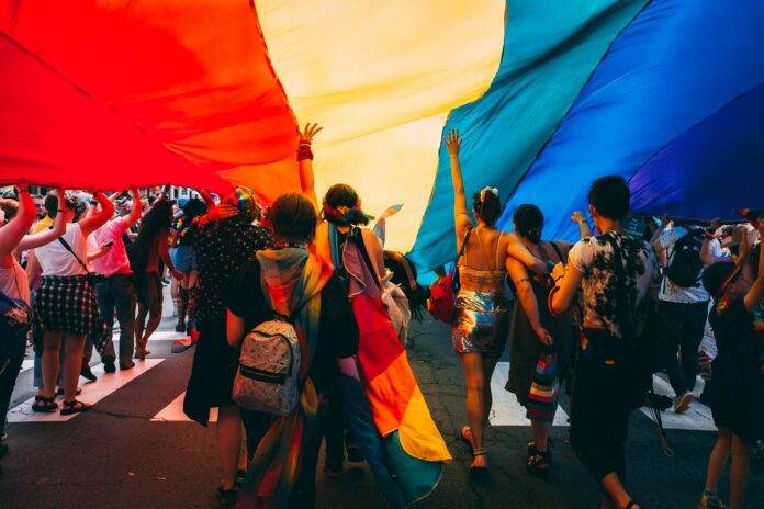 This is a photo of a pride parade. There are multiple people walking under a large rainbow flag. The camera is behind the people, so only their backs are visible.