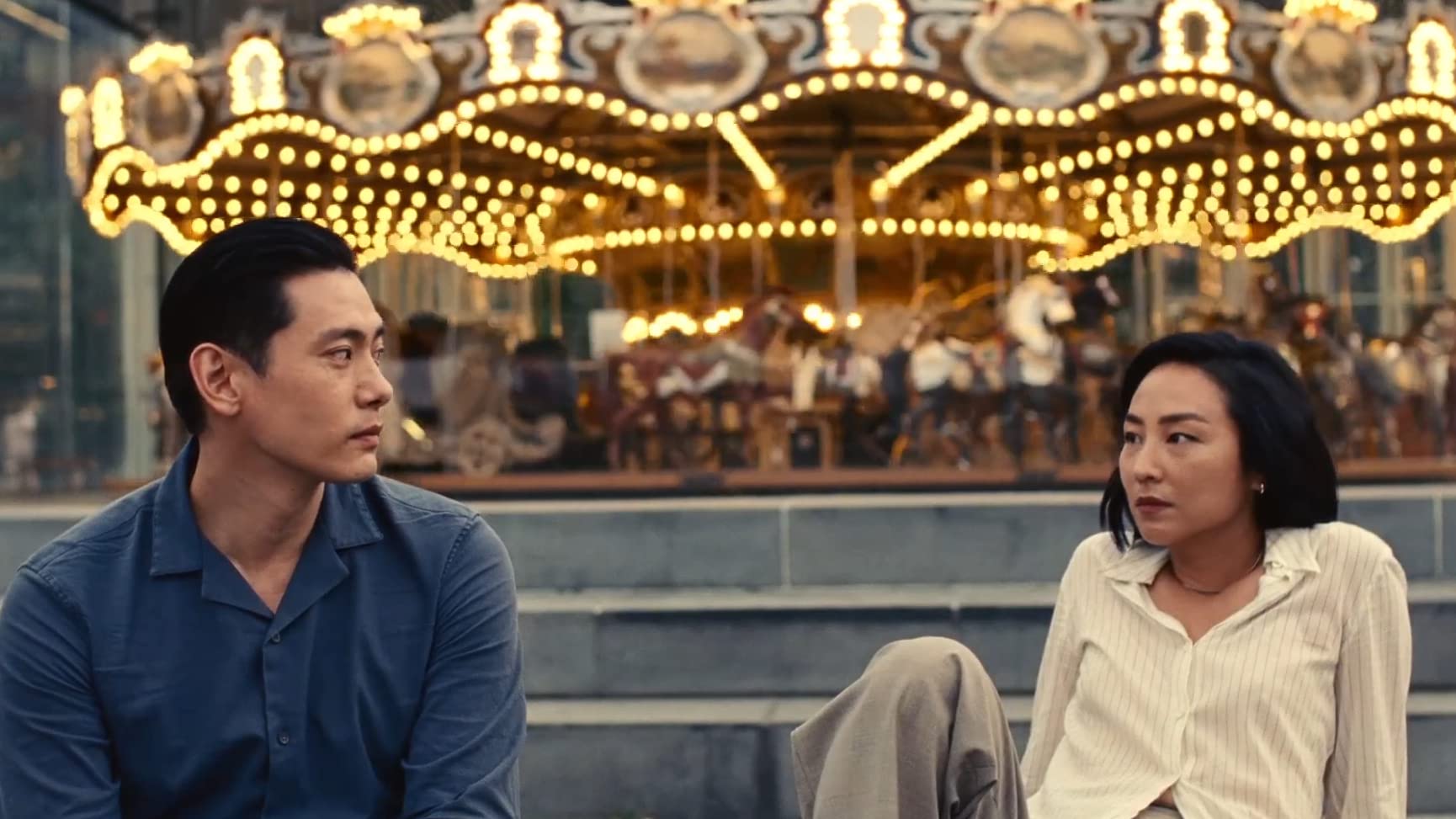 A Korean man and woman sit in front of a carousel blurred background with yellow lights. The woman is leaned back and they are looking at each other with subtle playful expressions.