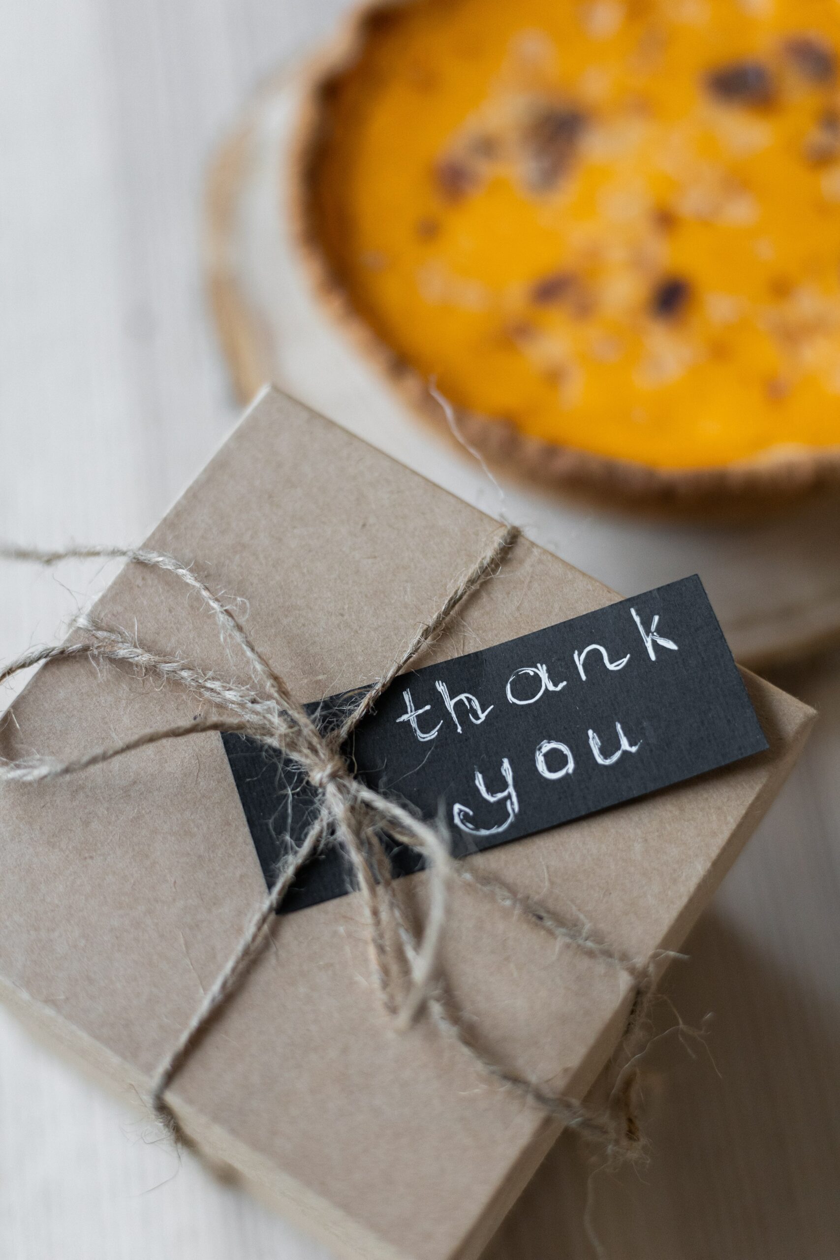 A photo of a small cardboard box wrapped in twine, which is woven into a bow at the top. It holds into place a small note with white text on black paper that writes “thank you.” An orange casserole dish is blurred in the background.