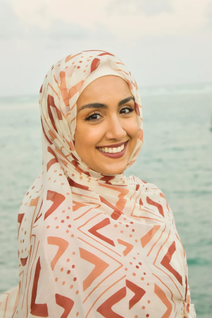 A headshot of Sumaiya Tufail smiling in front of an emerald ocean backdrop. She is wearing a pink head covering, and a dewy red lipstick.