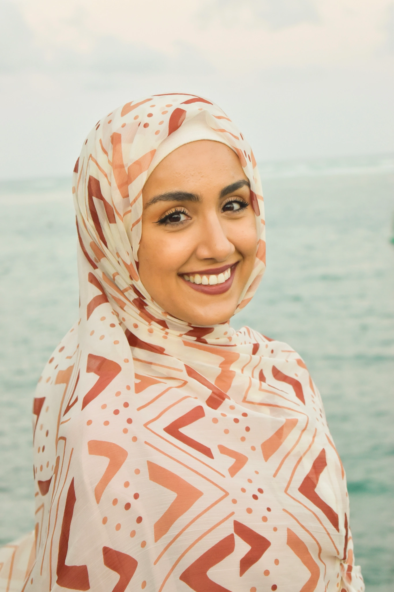 A headshot of Sumaiya Tufail smiling in front of an emerald ocean backdrop. She is wearing a pink head covering, and a dewy red lipstick.