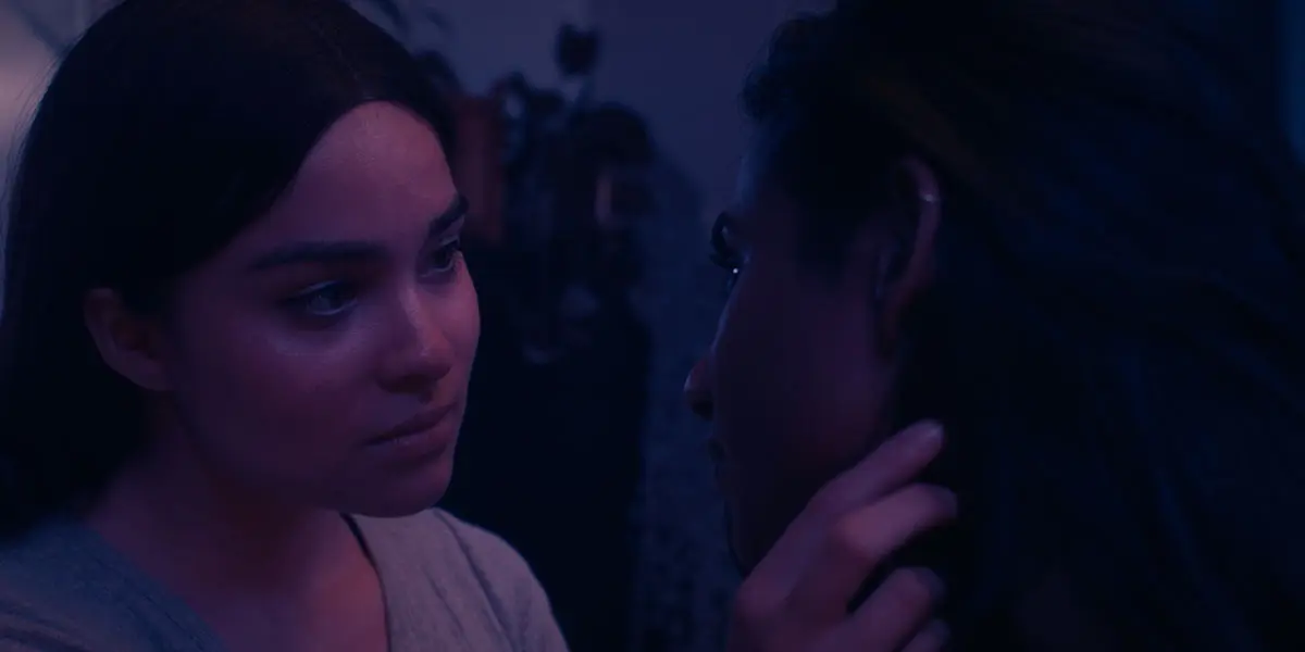 In dark, shadowy purple lighting, an intimate film still featuring a closeup of Kawenniióhstha gazing into Malai’s eyes with one hand on Malai’s chin. Both have long dark hair.