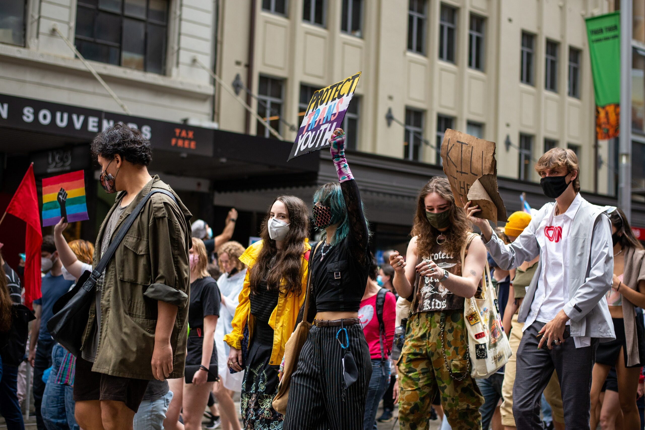 A group of people wearing masks and protesting, holding up a sign that says protect trans youth and pride flags.