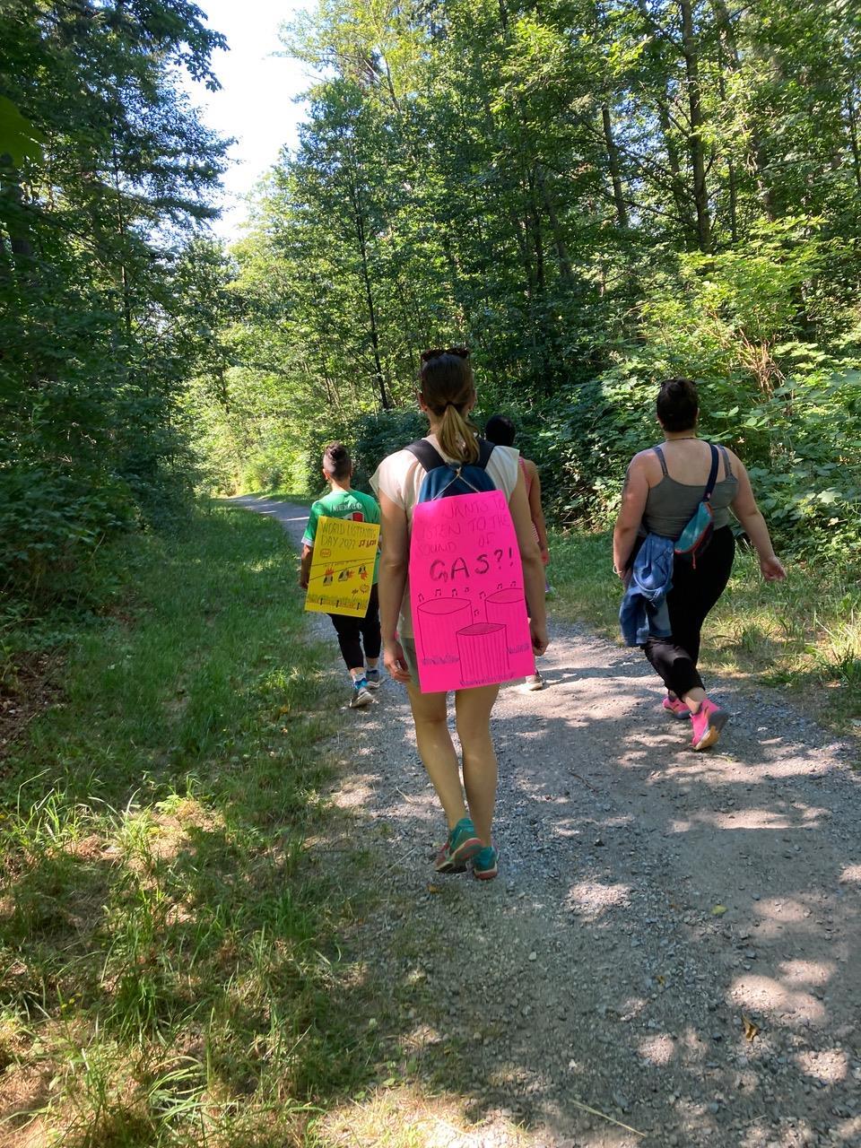 This is a photo taken on the World Listening Day Walk. People are walking down a Burnaby Mountain trail while wearing signs that read “Who wants to listen to the sound of gas” and “World Listening Day 2023.”