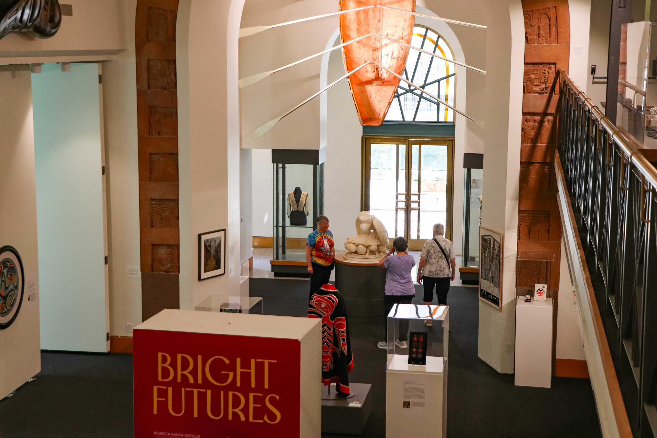 A photo taken from the mezzanine of a portion of the interior of the Bill Reid Gallery below. There is a bright salmon-coloured canoe with rows hanging from the ceiling, illuminated by sunlight through the windows. A large board in the middle of the space that read “Bright Futures.” There are various artworks behind glass and people are walking around, looking.