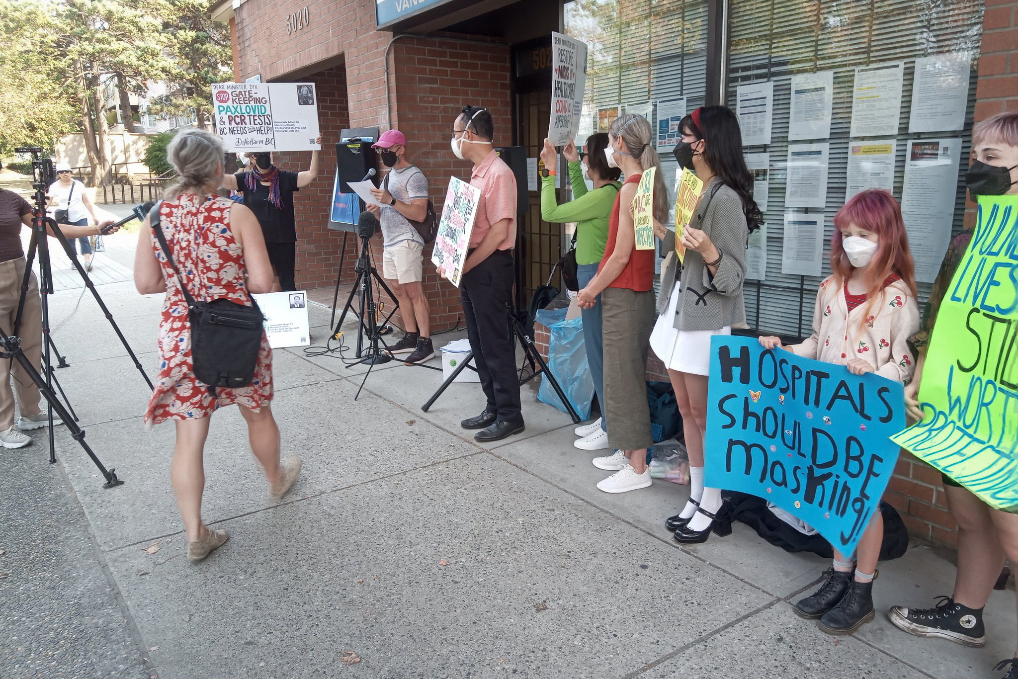 News crews congregate outside Adrian Dix's MLA office, gathering around protesters wearing respirator masks and holding signs demanding masks back in healthcare, access to Paxlovid and PCR testing, and holding some of the names of those impacted. In the centre, a young person stands at a microphone telling her story. She holds a yellow sign saying she has had Long Covid for 20+ months.