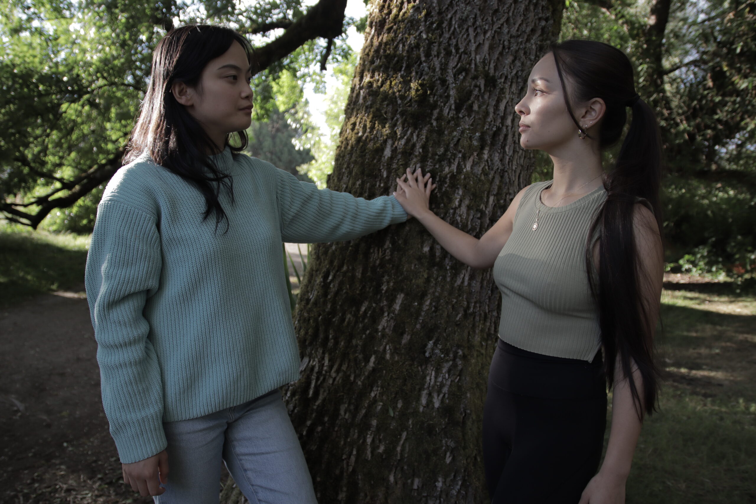 Two Asian women look at each other with their hands touching on a tree branch between them. They’re both wearing neutral colored shirts and they’re in a forest surrounded by trees.