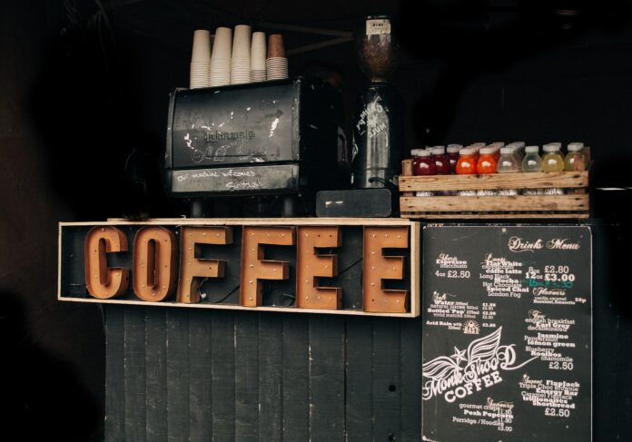 Café counter with a large sign that says “coffee.” The espresso machine’s paint is peeling and there’s “our machine welcomes scribbles!” scratched onto it.