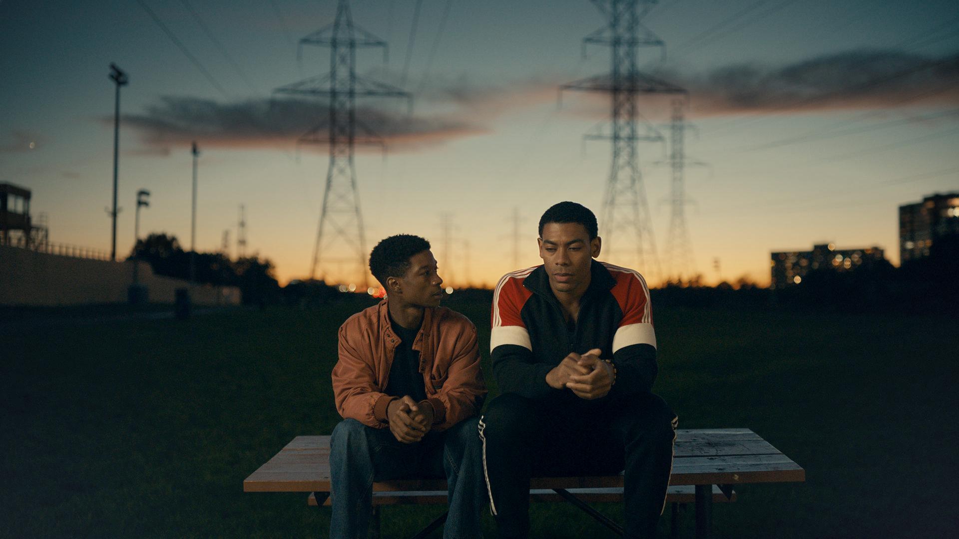 Two brothers, Black young men, sit on a bench outside in front of power lines. The sun is setting and the sky is tinted orange.
