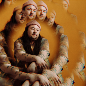 Prisms by G. R. Gritt features Gritt in an earthy tie dye shirt and beanie with an orange, fume-y atmosphere surrounding them.