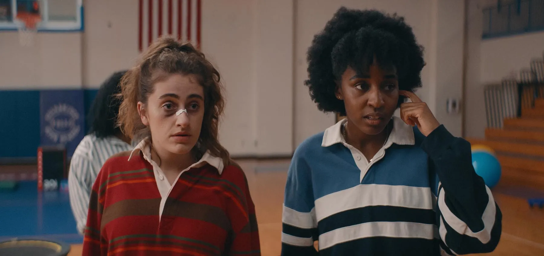 A film still featuring two protagonists of Bottoms, PJ and Josie, in a school gym looking to the side, looking bewildered. PJ has a slight black eye and a bandaid on her nose.