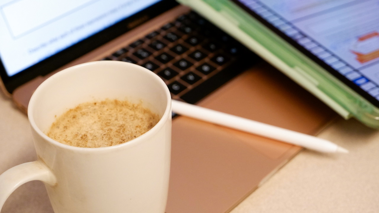 An coffee mug held in front of someone’s desk with laptop and study material laid out on it.