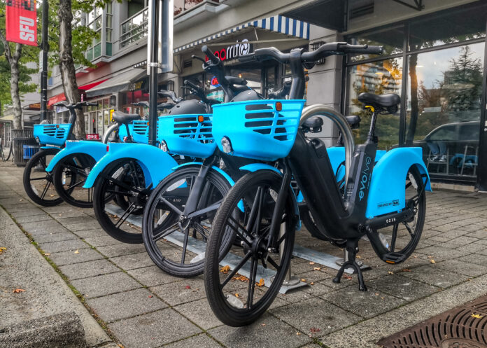 This is a photo of the blue Evolve E-Bikes outside of the SFU Burnaby campus. A row of Bikes are lined up. The bikes have baskets in the front where helmets and safety information sit.