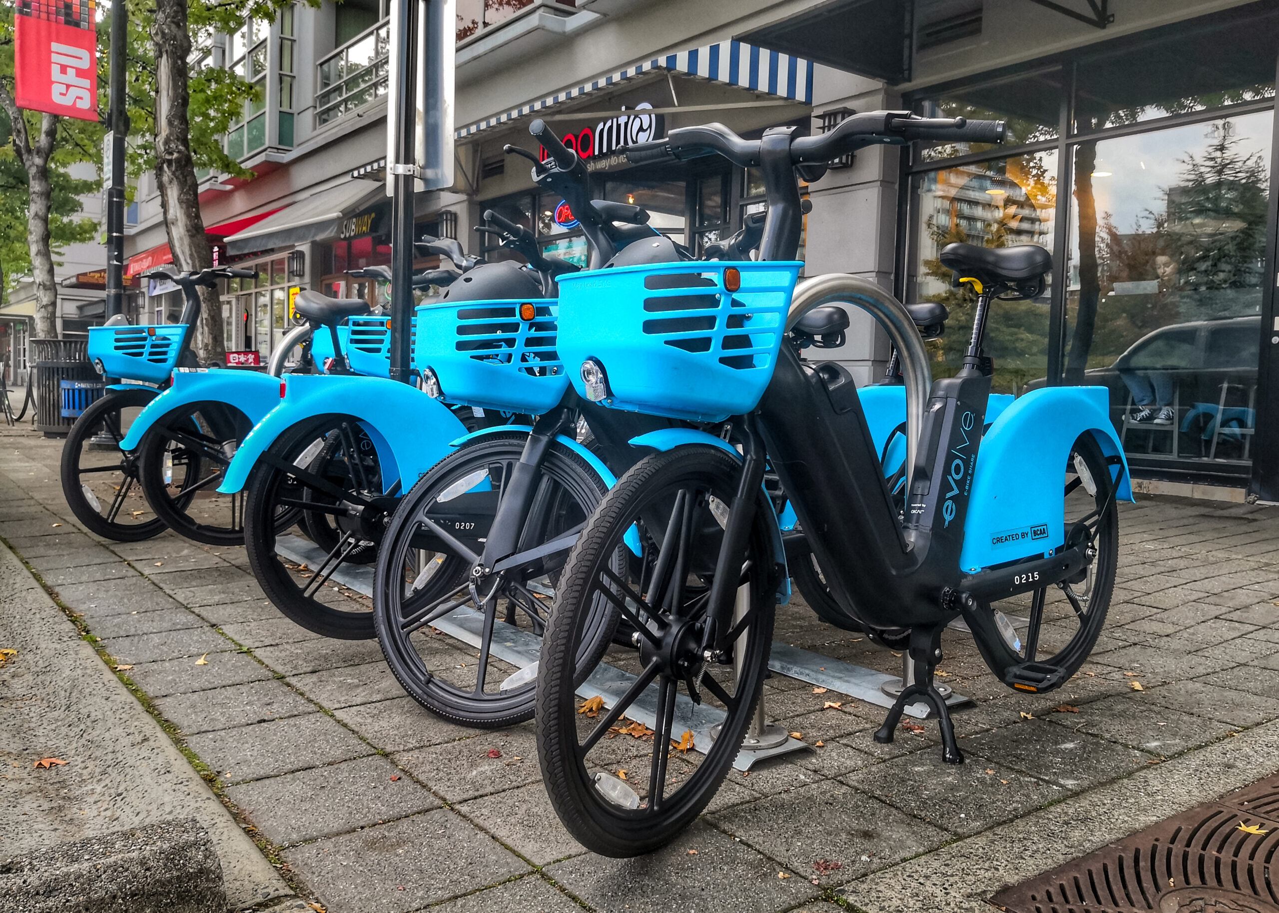 This is a photo of the blue Evolve E-Bikes outside of the SFU Burnaby campus. A row of Bikes are lined up. The bikes have baskets in the front where helmets and safety information sit.