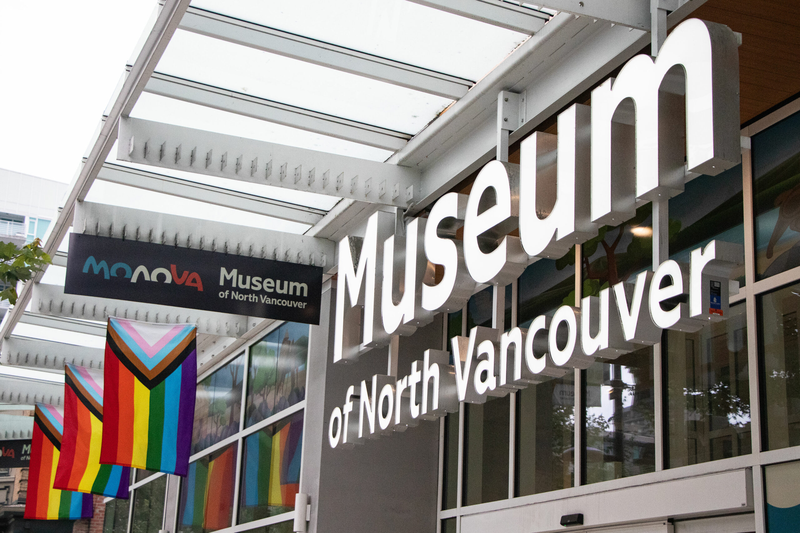 Exterior of building with a closeup of the words “Museum of North Vancouver” in bold white block letters in front a window. There are trans flags and pride flags hanging from the canopy.