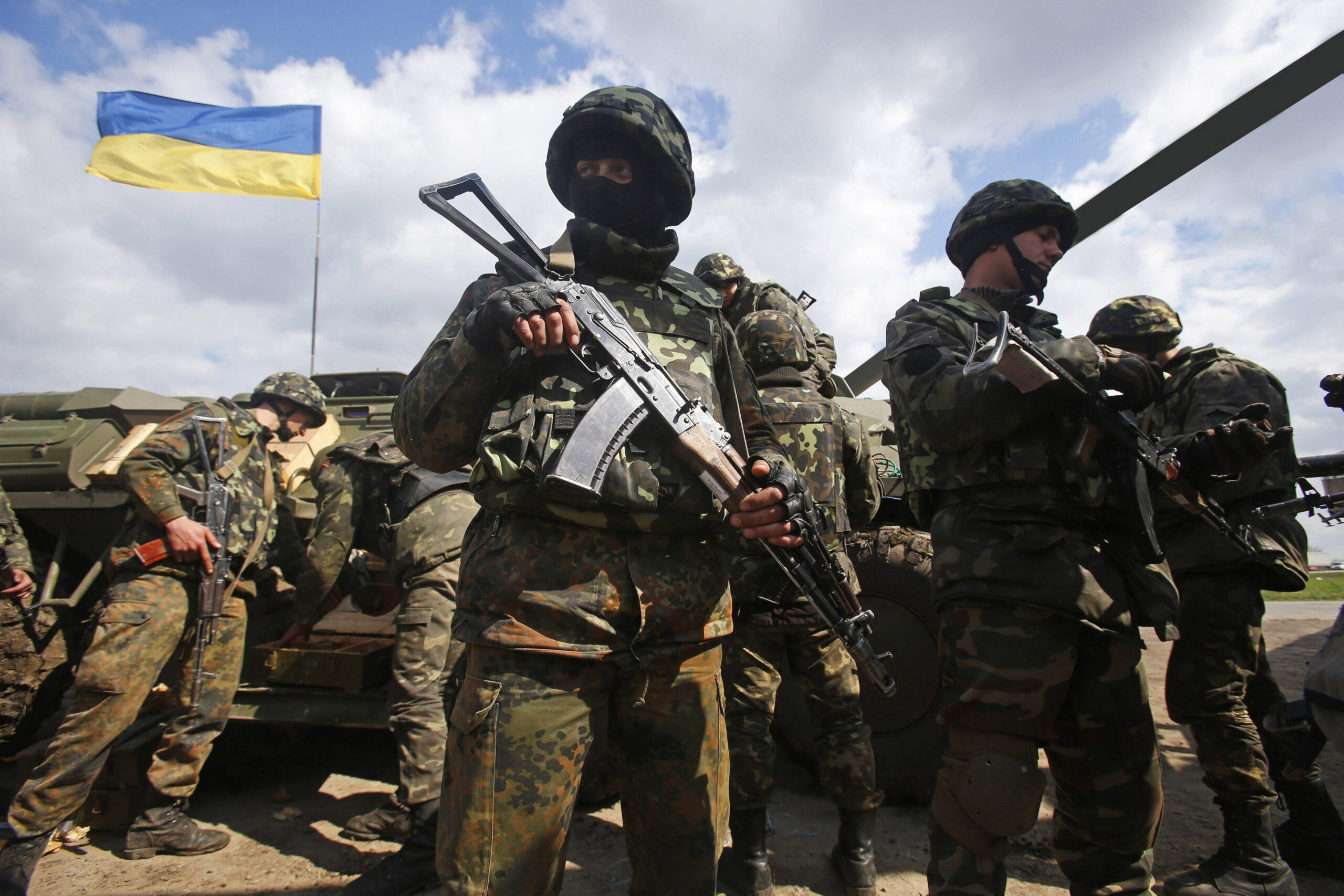 This is a photo of Ukrainian soldiers. They are standing in front of a Ukrainian flag, as it is waving. The soldiers are dressed in military gear, and are holding weapons.