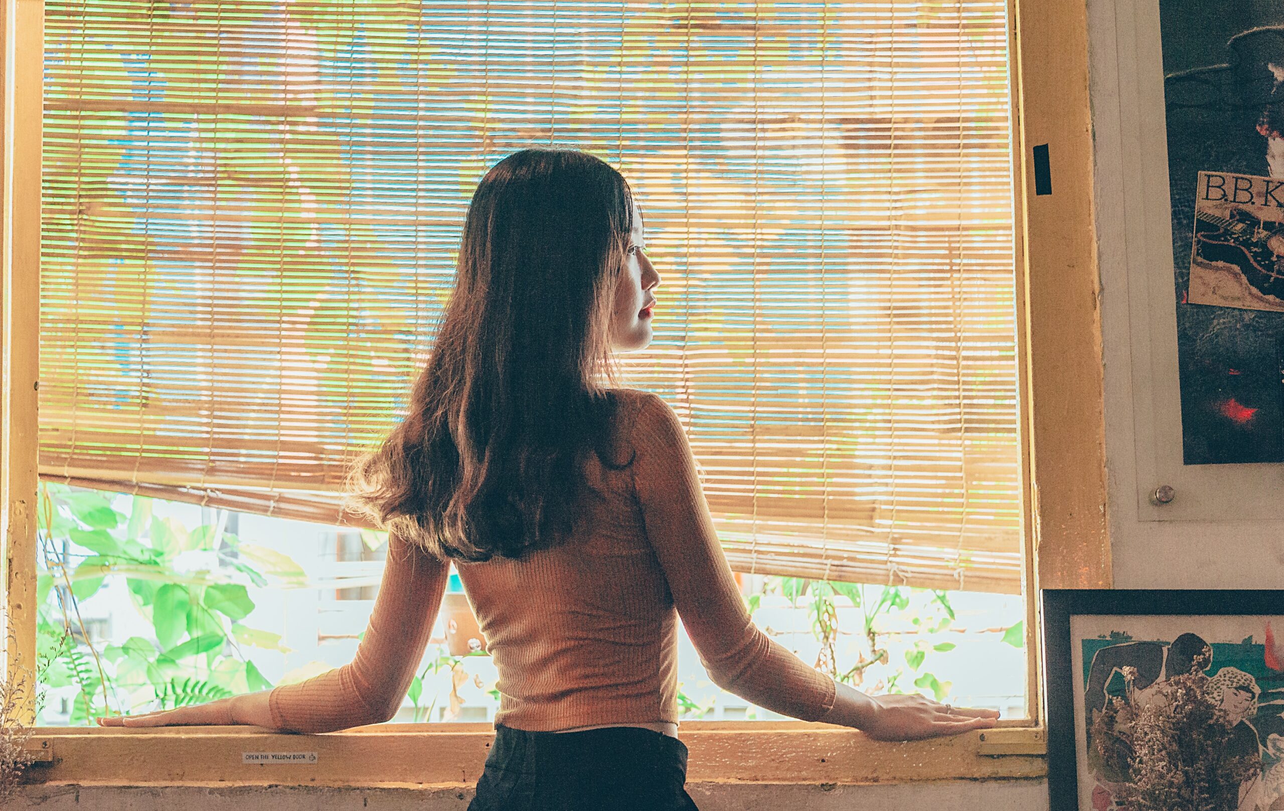 A woman standing beside a window. The blinds are slightly raised.