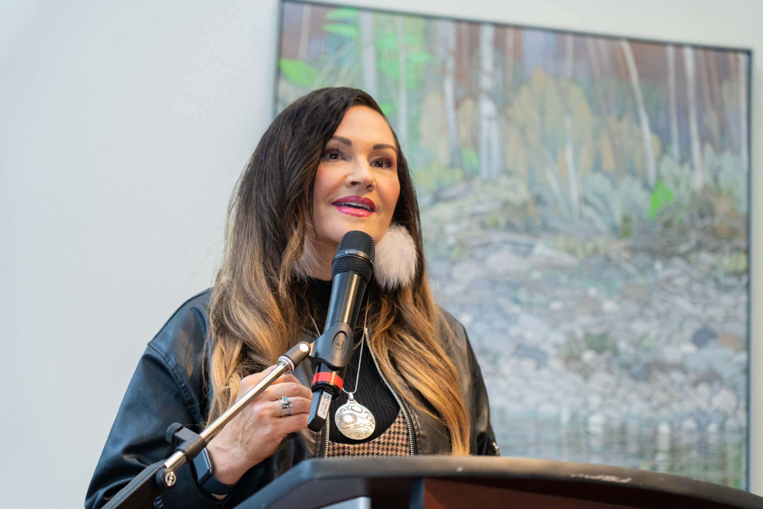 This is a photo of Angela Sterritt giving her lecture. She is standing at a podium, speaking to a large audience. The photo is close on her face. Behind her there is a painting of nature, forest, and animals.