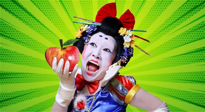 GUMBO A Japanese person in Kabuki-style pale white foundation with exaggerated black and red makeup, hair up in a bow with chopsticks and flowers adorning their hair, holding a red apple. They are behind a green, comic-book style background with an exaggerated facial expression, white gloves, and a fairytale-style blue dress.