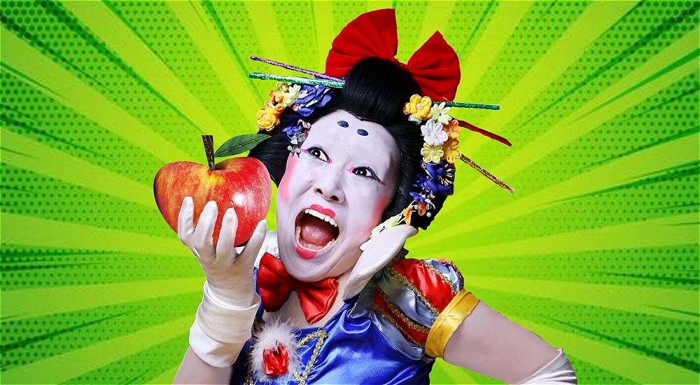 A Japanese person in Kabuki-style pale white foundation with exaggerated black and red makeup, hair up in a bow with chopsticks and flowers adorning their hair, holding a red apple. They are behind a green, comic-book style background with an exaggerated facial expression, white gloves, and a fairytale-style blue dress.