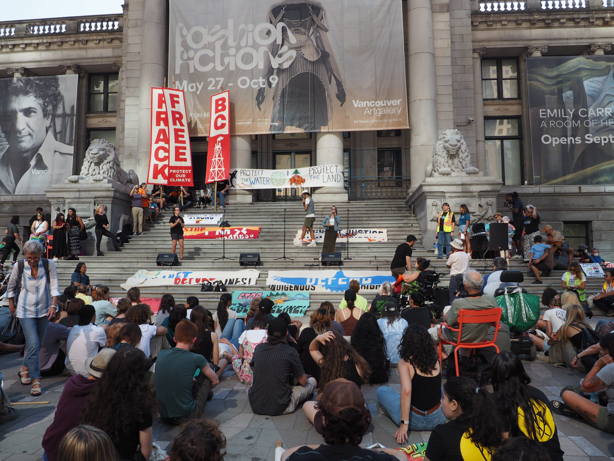 This is a photo of the strike, being held outside the Vancouver Art Gallery. Numerous people are gathered outside the gallery, holding signs that show support for the climate protest. The largest signs, placed behind the speakers, read “Frack Free BC.”