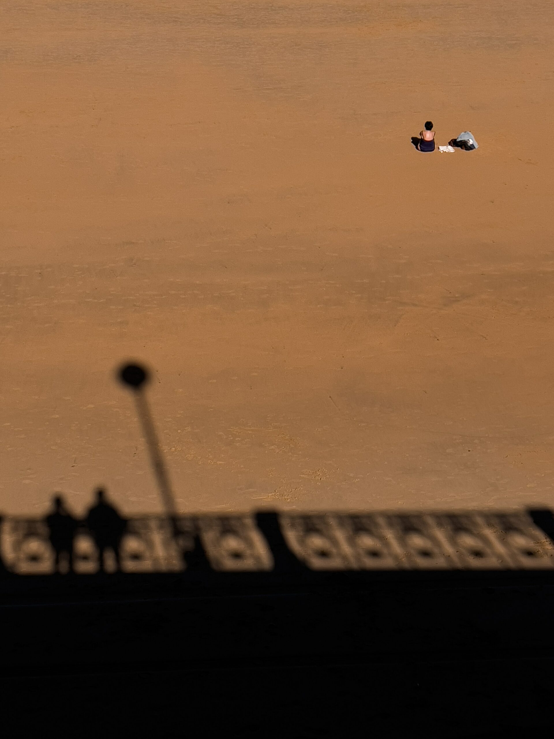 A person sitting alone on a large expanse of a sandy beach. Behind them is a shadow of two people standing together on a bridge.