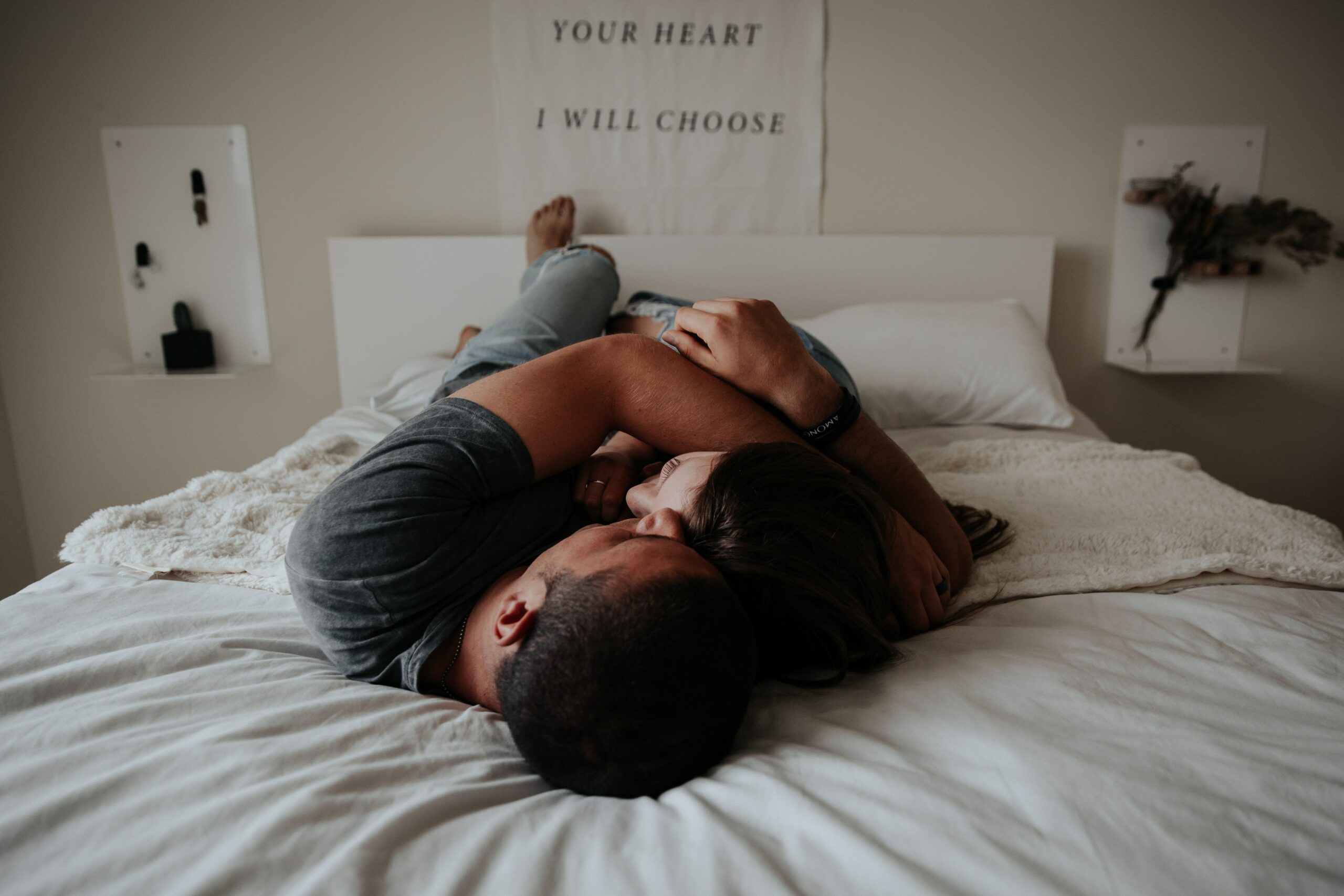 This is a photo of a couple hugging, while lying down on their bed. They are wrapped in eachothers arms closely.