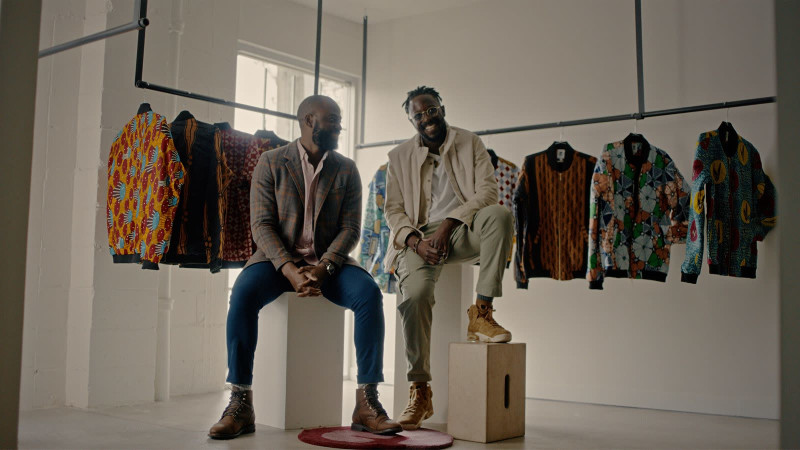 Two young Black men sitting casually and smiling in front of a display of garments hanging on a rack, with colourful patterns.