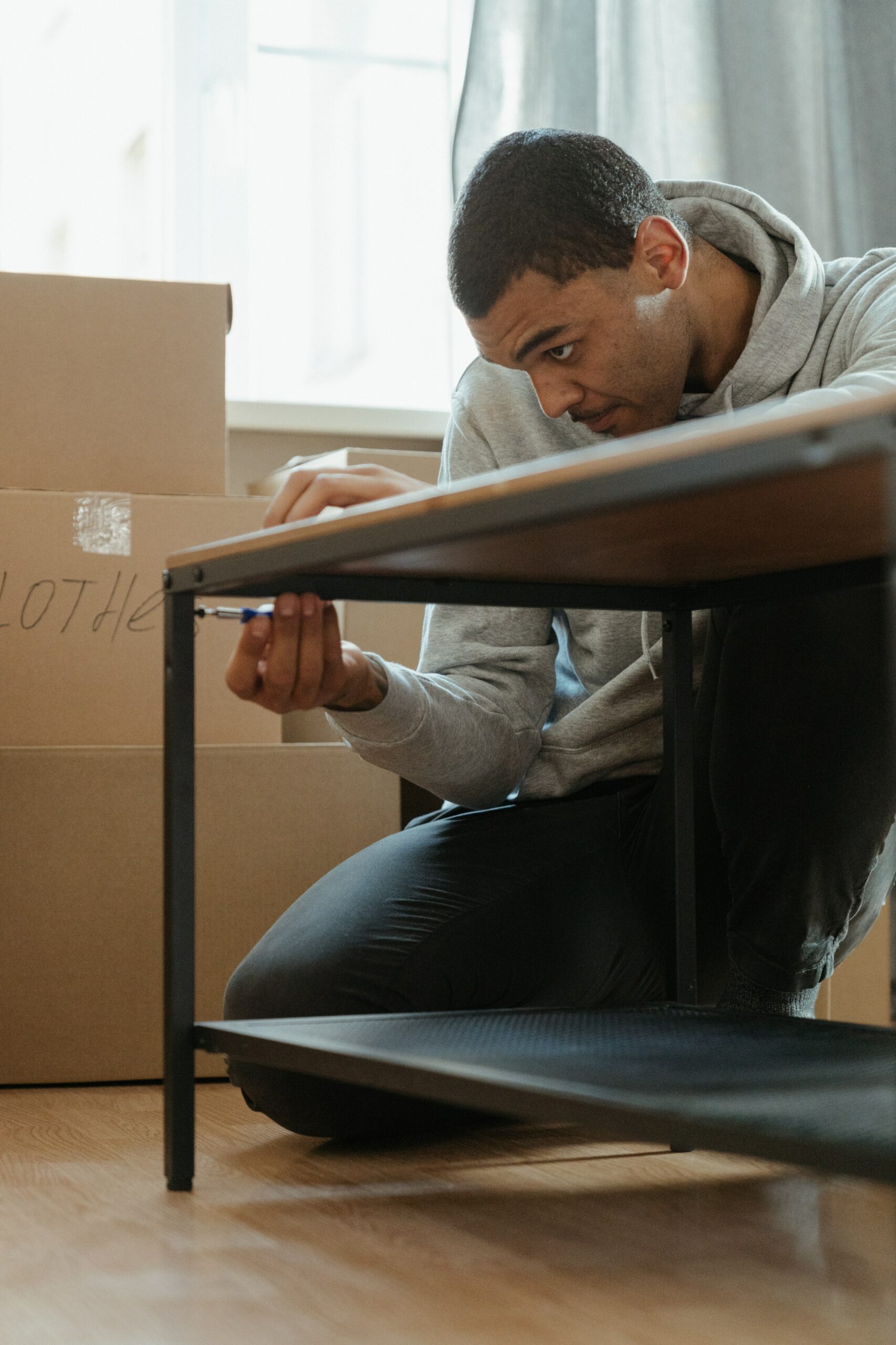 Man wearing a grey hoodie surrounded by cardboard boxes. He is tightening a screw as a part of assembling a table.