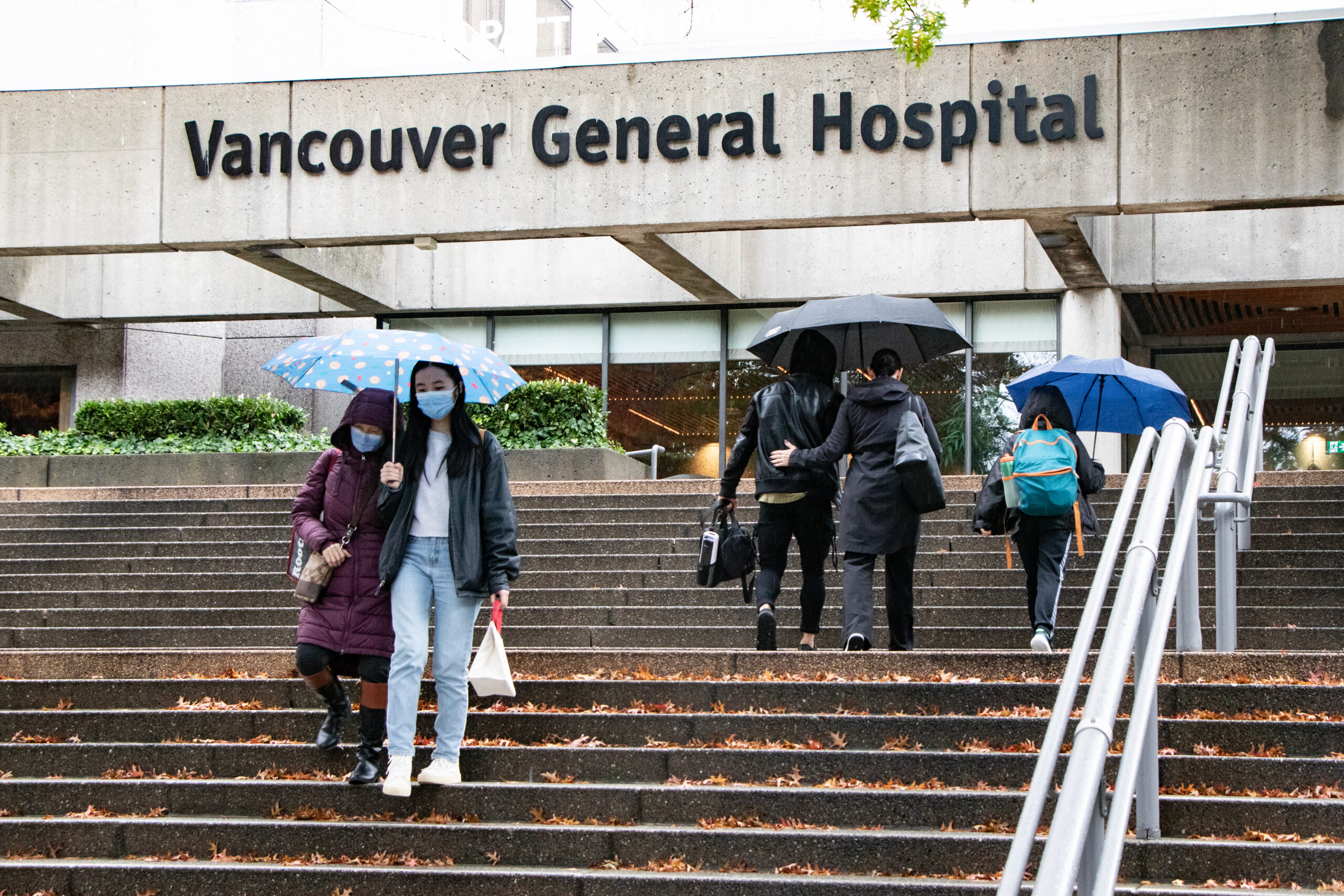 This is a photo of the outside of Vancouver General Hospital. As it is raining outside, there are people walking in and out of the building holding umbrellas.